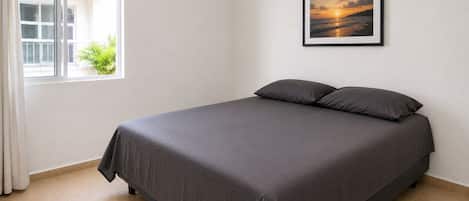 Una cama bien hecha con una colcha oscura y almohadas en una habitación con un ventanal grande y un cuadro en la pared.