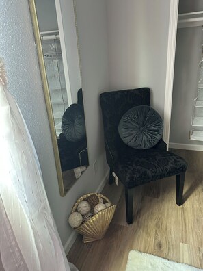 A room with a dark-colored chair, a gold-framed mirror, and a basket with decorative items.