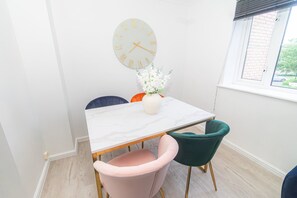 Ein Essbereich mit einem weißen Tisch, zwei Stühlen, einer Vase mit weißen Blumen und einer Wanduhr.