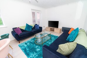 Ein modernes Wohnzimmer mit einer Sofagarnitur, einem Glastisch, einem Flachbildfernseher und einem Fenster mit Jalousien.