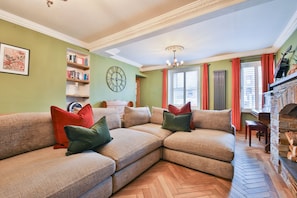 A living room with a sectional sofa, a clock, a fireplace, and a bookshelf. - The Swansea Homestay | Spacious 4-Bed Retreat (Swansea)