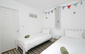 A room with two single beds, a white door, a small bedside table, a mirror, and a striped carpet. - Atlantic Reach – Cosy 2-Bed with Parking (Newquay)