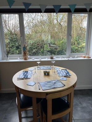 A round wooden table with a white surface, surrounded by black cushioned chairs, positioned in front of a window with a view of trees and a bird feeder. - Atlantic Reach – Cosy 2-Bed with Parking (Newquay)