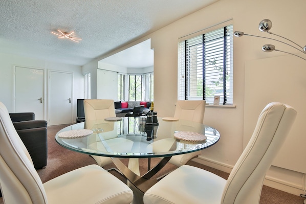 A modern living room with a glass table and white leather chairs, a sofa with cushions, and a television. - Swansea Waterfront 1 Bed Apt with Parking (Swansea)
