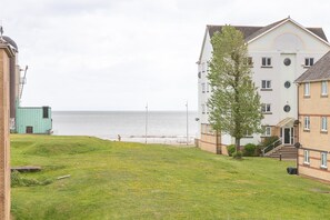 A coastal residential area with a grassy lawn, a building with multiple windows, and a view of the sea. - Swansea Waterfront 1 Bed Apt with Parking (Swansea)