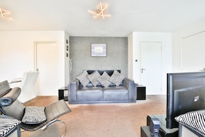 A modern living room with a grey sofa, patterned cushions, a flat-screen TV, and a unique ceiling light fixture. - Swansea Waterfront 1 Bed Apt with Parking (Swansea)