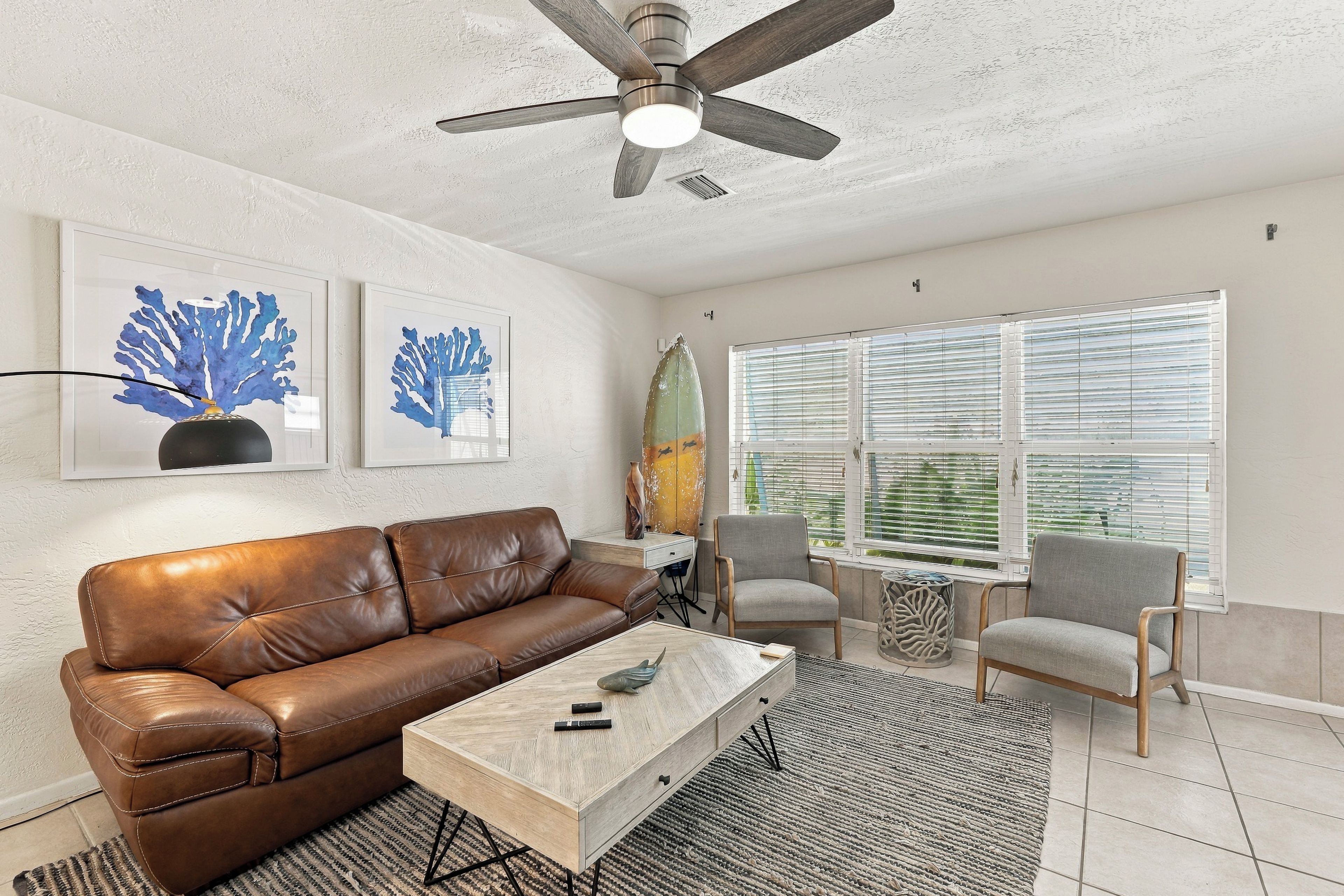 A living room with a brown leather sofa, a wooden coffee table, two chairs, a ceiling fan, and artwork on the wall.