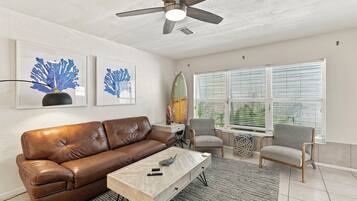 A living room with a brown leather sofa, a wooden coffee table, two chairs, a ceiling fan, and artwork on the wall.