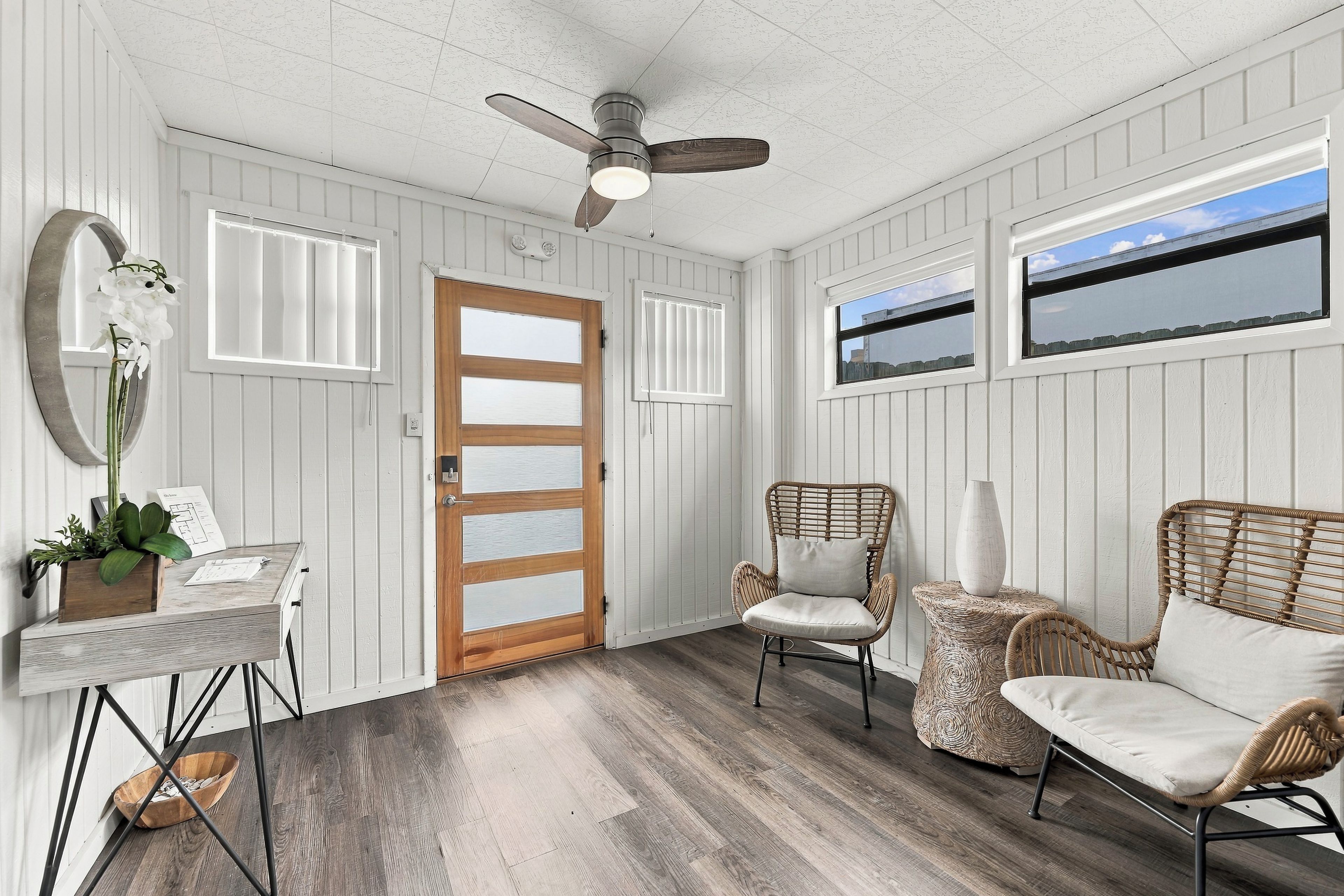 A room with a ceiling fan, a wooden door, two windows, wicker furniture, and a small table with a plant.