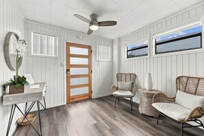 Une pièce dotée d’un ventilateur de plafond, d’une porte en bois, de deux fenêtres, de meubles en osier et d’une petite table sur laquelle est posée une plante.