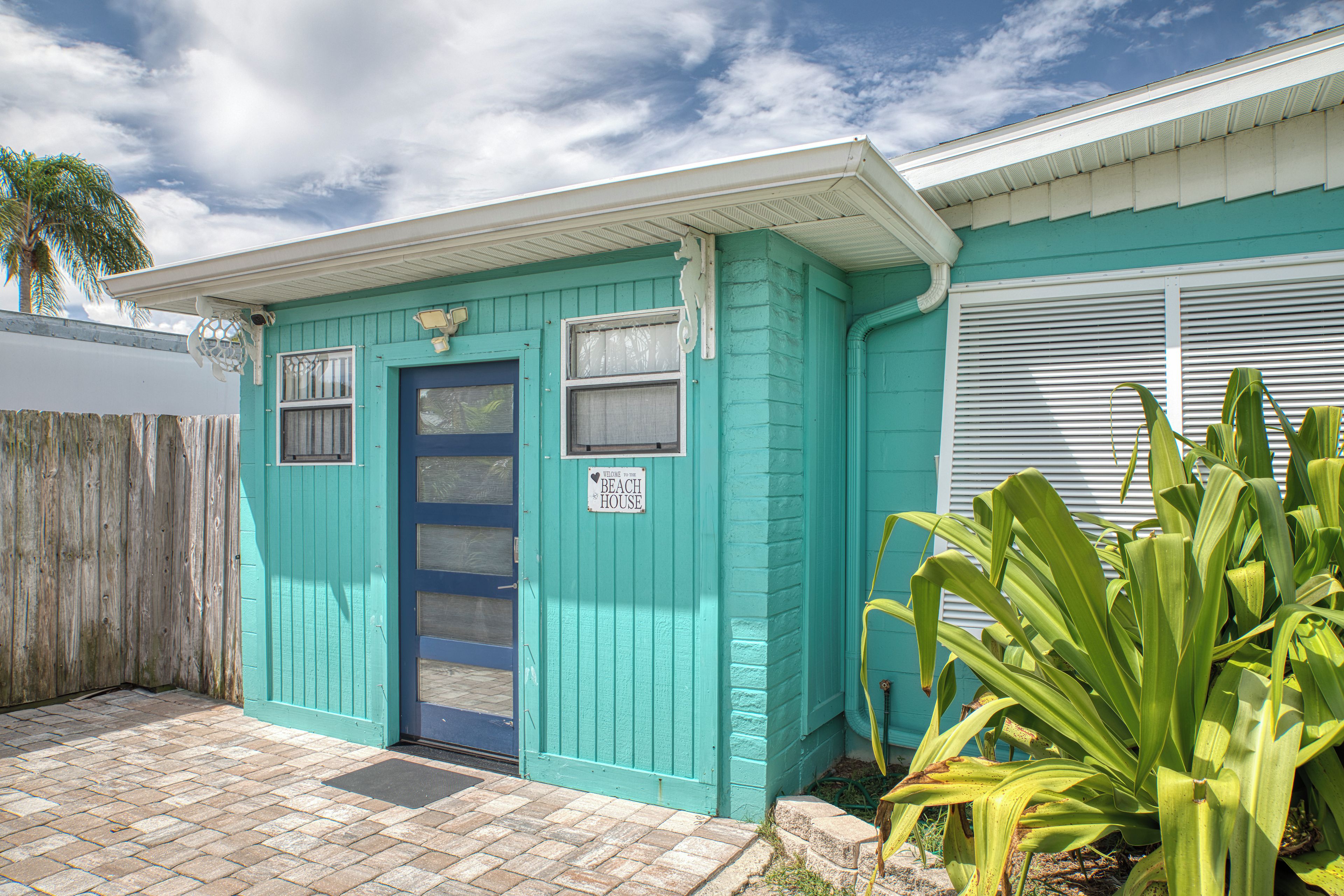 A small, single-story building with a blue door and windows, a sign reading 'Welcome to the Beach House,' and a plant in the foreground.