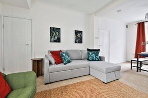 Ein modernes Wohnzimmer mit einem grauen Ecksofa, einem grünen Sessel und einem braunen Teppich. An der Wand hängen zwei gerahmte Kunstwerke, eine weiße Tür und ein roter Vorhang am Fenster.
