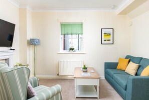 A living room with a sofa, a coffee table, a television, a radiator, a window with blinds, and a framed picture on the wall. - Seaview 2-Bed with Private Parking, Portishead (Portishead)