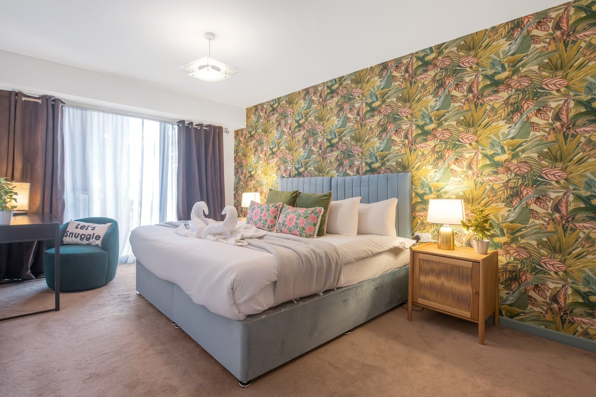 A hotel room with a bed, a desk, a chair, and a wall with a floral pattern.