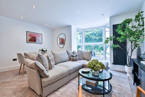A modern living room with a sofa, a round glass coffee table, and a large window offering a view of greenery. - Central 4-Bed Apartment on Mumbles Road (Swansea)