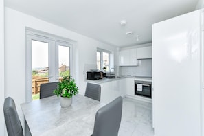 A modern kitchen with white cabinets, a dining area with a table and chairs, and a view of the outside through large windows. - Modern 2-Bed Home w/ EV Charger & Garden (Nottingham)