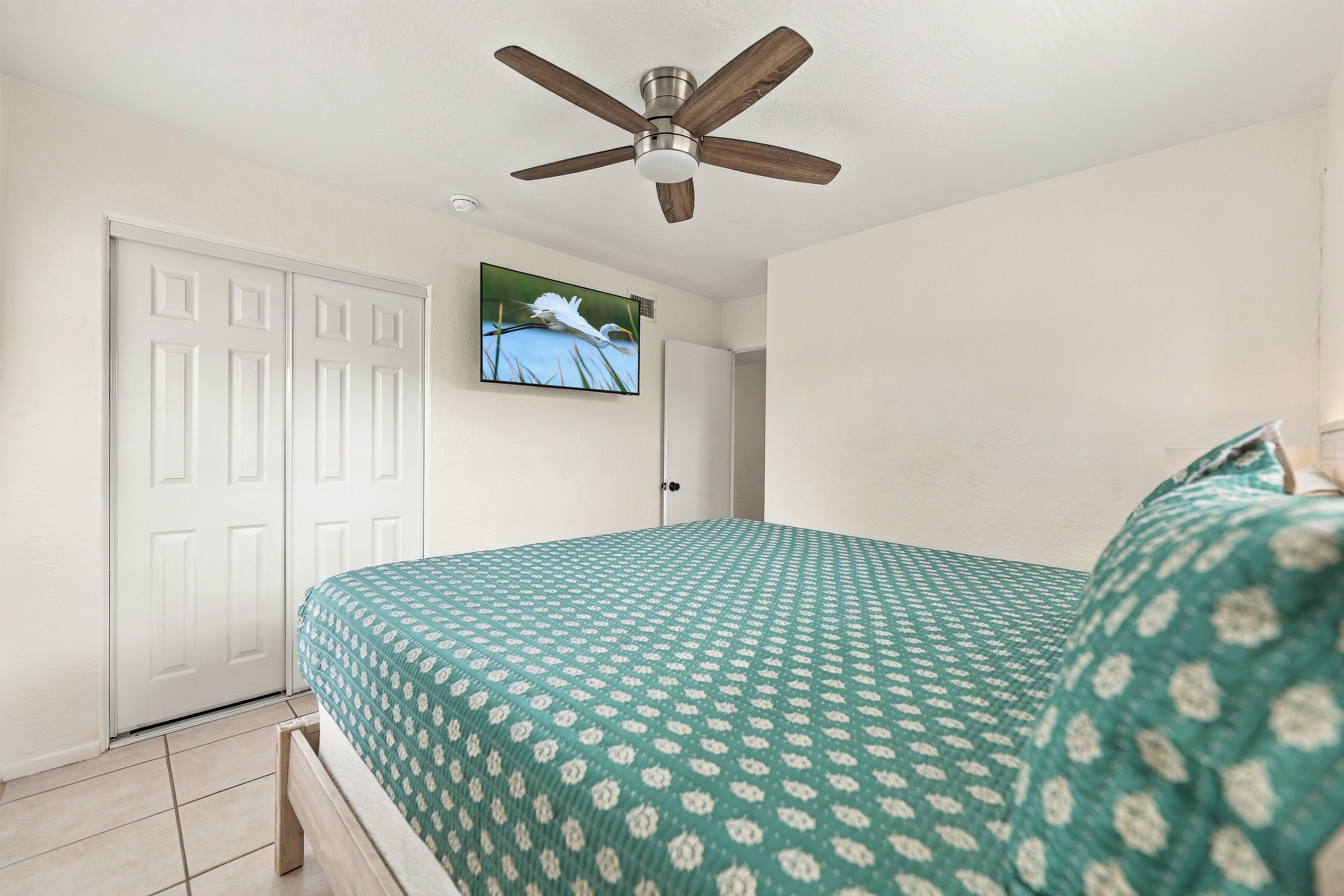 Une chambre à coucher avec un lit, un ventilateur de plafond, un téléviseur fixé au mur et une porte blanche.
