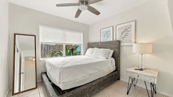 Une chambre à coucher comprenant un lit, une table de chevet, une lampe, un ventilateur de plafond, une fenêtre avec des stores et des œuvres d’art encadrées accrochées au mur.