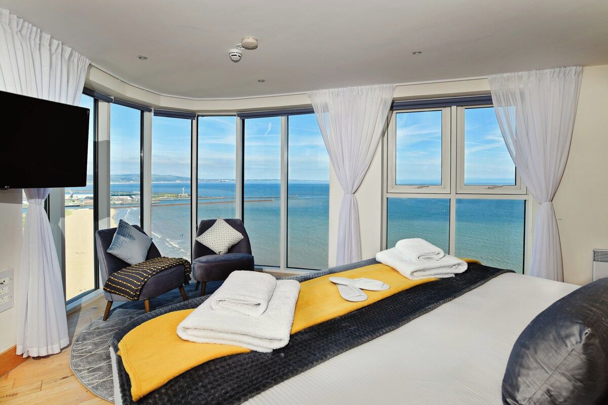 A hotel room with a large bed, a TV, a seating area with a view of the sea, and a balcony.