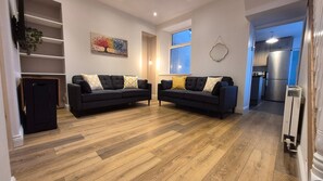 A living room with two sofas, a painting, a window, and a kitchen area. - Swansea City & Bay 4-Bedroom House (Swansea)