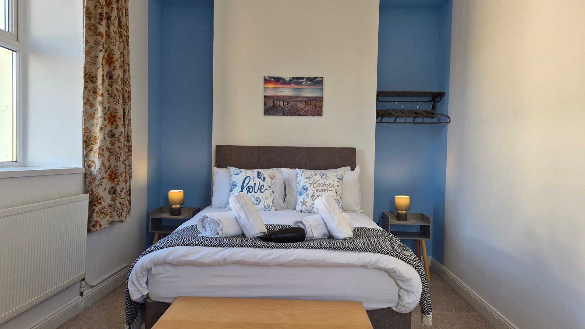 Un dormitorio con una cama, dos mesitas de noche, un mueble auxiliar, una ventana, una cortina y un estante montado en la pared.