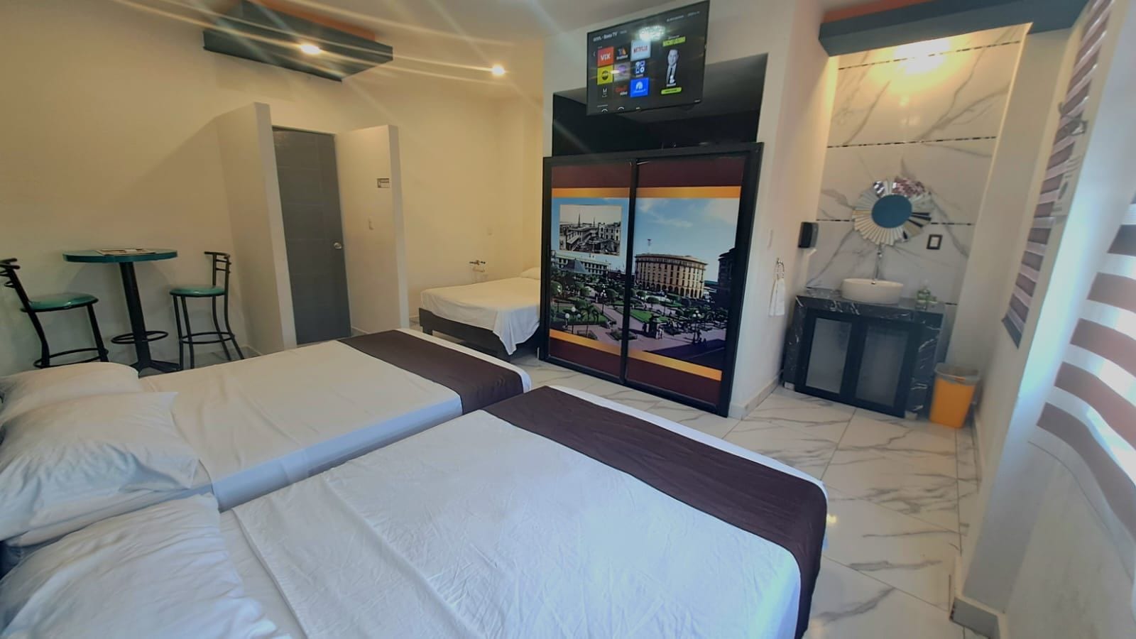 A hotel room with two beds, a TV, a small table with chairs, and a view of a cityscape through a glass door.