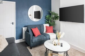Ein Wohnzimmer mit blauer Wand, einem grauen Sofa mit roten Kissen, einem runden Spiegel, einem Flachbildfernseher, einem weißen Couchtisch mit Dekoration und einer Topfpflanze.