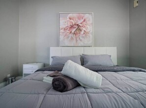 Un dormitorio con una cama, almohadas, una mesita de noche y una obra de arte colgada en la pared.