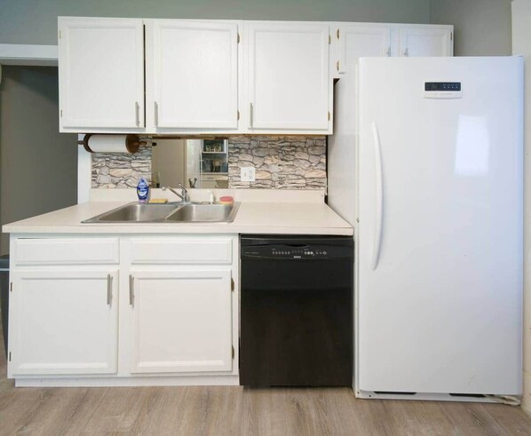 Una cocina con armarios blancos, protector contra salpicaduras de piedra, lavavajillas y refrigerador blanco.