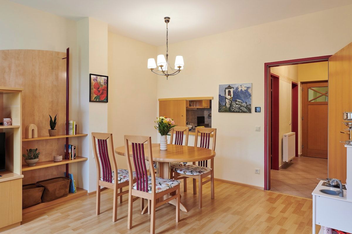 Un comedor con mesa y sillas de madera, una araña y una cocina al fondo.