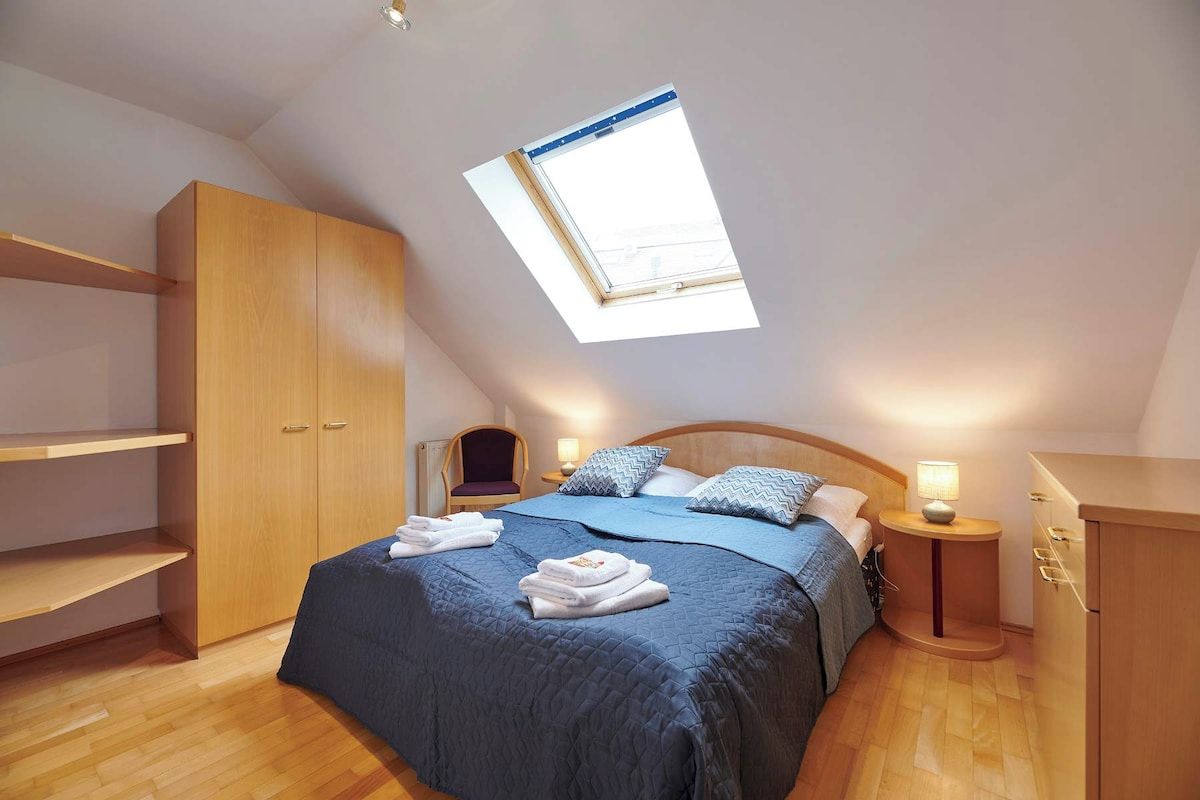 Ein Schlafzimmer mit Dachfenster, einem Bett mit blauer Bettwäsche, Holzmöbeln und einem kleinen Nachttisch mit Lampe.