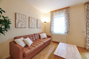 Una sala de estar con un sofá de cuero marrón, una mesa de centro de madera y un radiador. Hay tres cuadros colgados en la pared y un ventanal con cortinas florales.