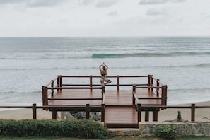 A wooden deck overlooking a beach with waves, a person meditating, and a stone wall in the foreground. - Casa Privada Frente al Mar | Lujo, Playa y Exclusi (Troncones)