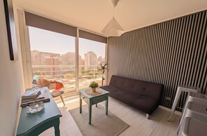 A modern living room with a sofa, a small table with a plant, and a view of the city through large windows. - Torre San Isidro by Domus Travels (Santiago)