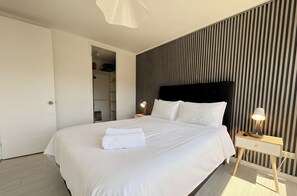 A neatly made bed with white linens and a folded towel, a bedside table with a lamp, and a wardrobe with a mirror. - Torre San Isidro by Domus Travels (Santiago)