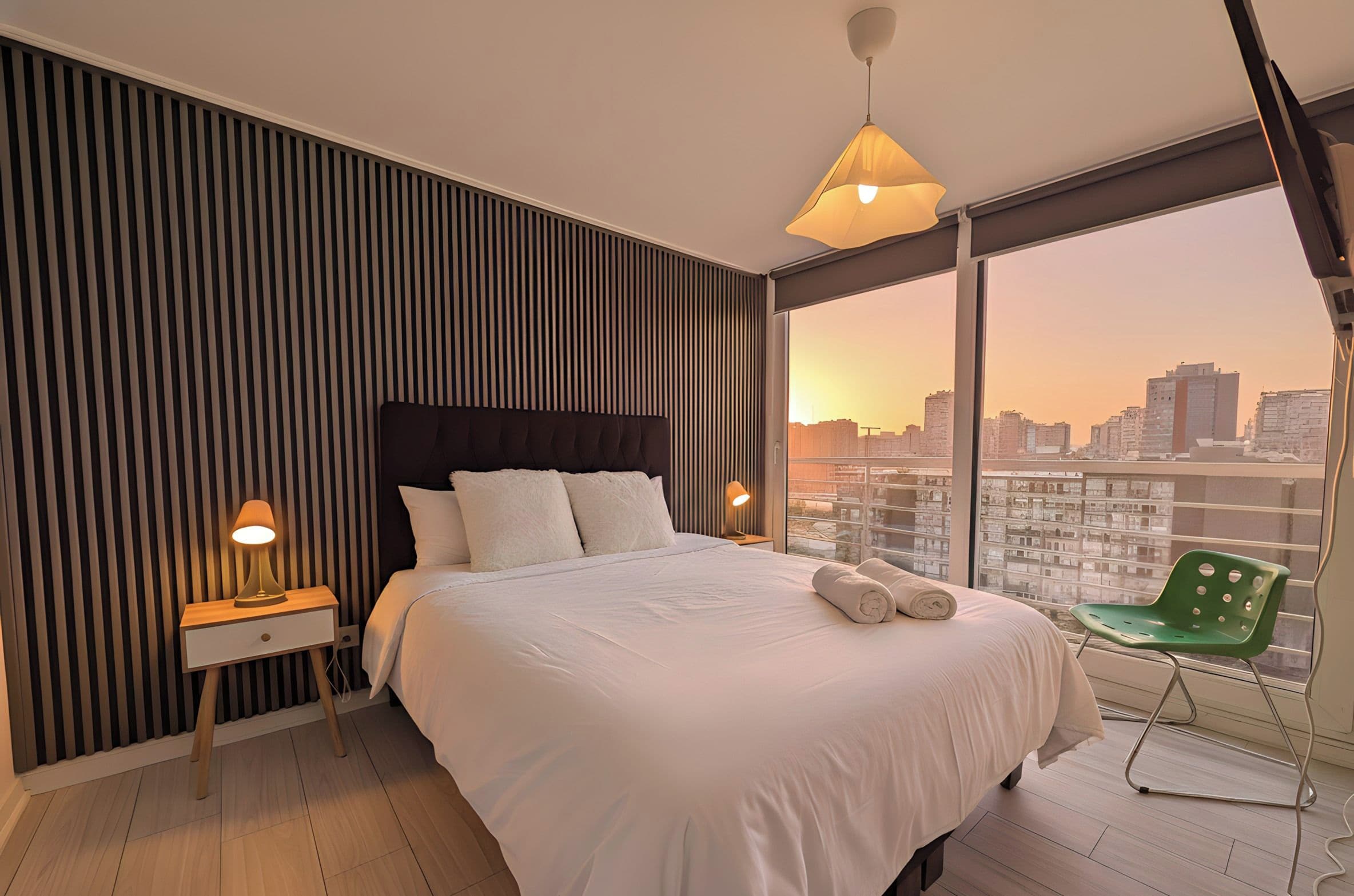 A modern bedroom with a large bed, a nightstand, a lamp, a green chair, and a city view through large windows.
