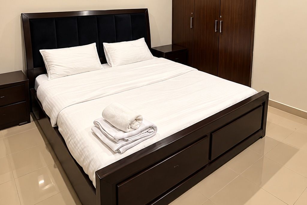 Een net opgemaakt bed met witte lakens en een opgevouwen handdoek, een donker houten hoofdbord en een houten garderobe met twee deuren.