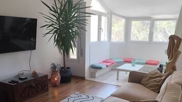 Una sala moderna con sofá, televisión, planta en maceta y zona de estar con cojines.