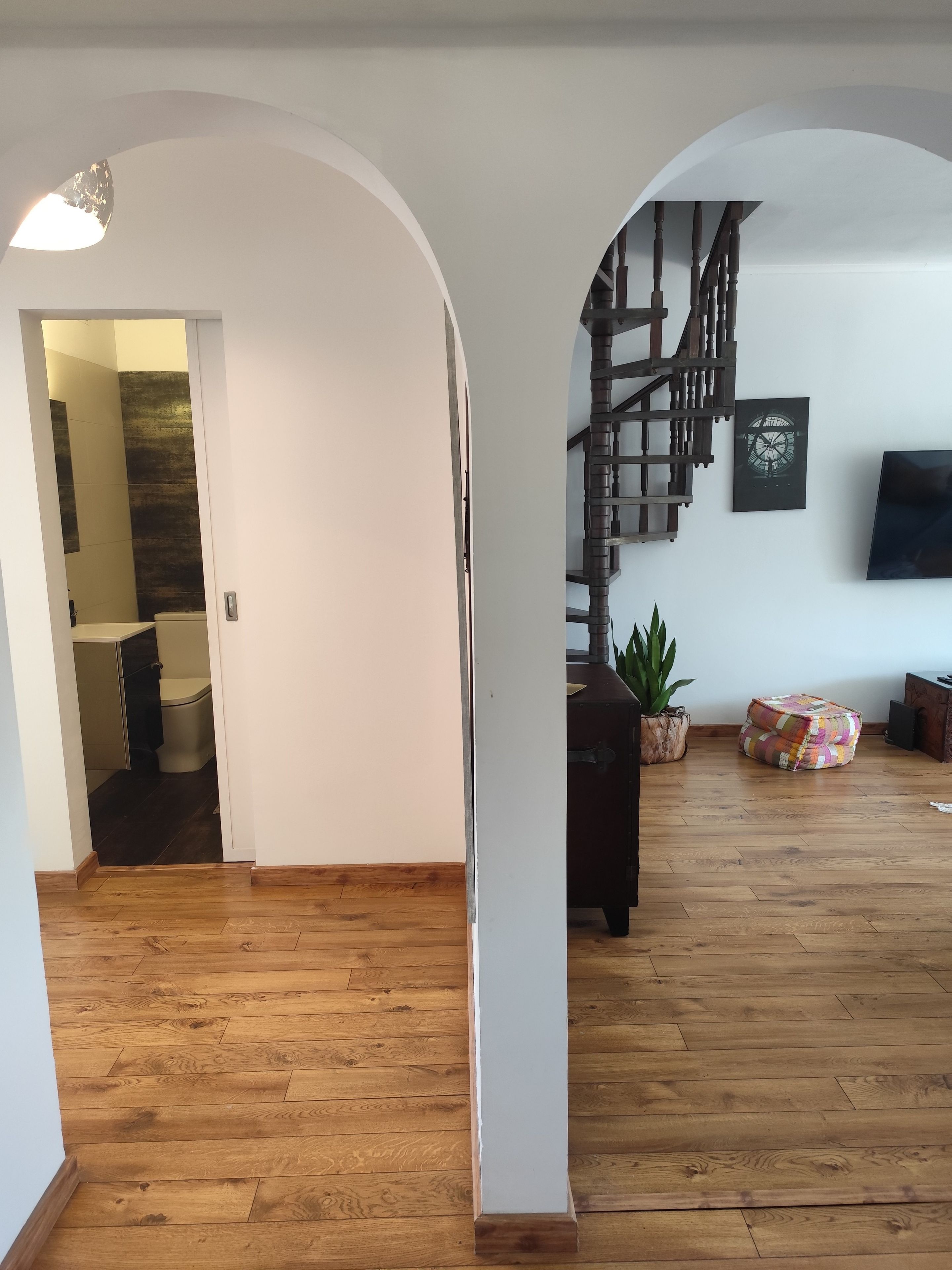Un espacio habitable amplio con piso de madera, una escalera con barandilla de madera, un televisor de pantalla plana en la pared y una planta en maceta.