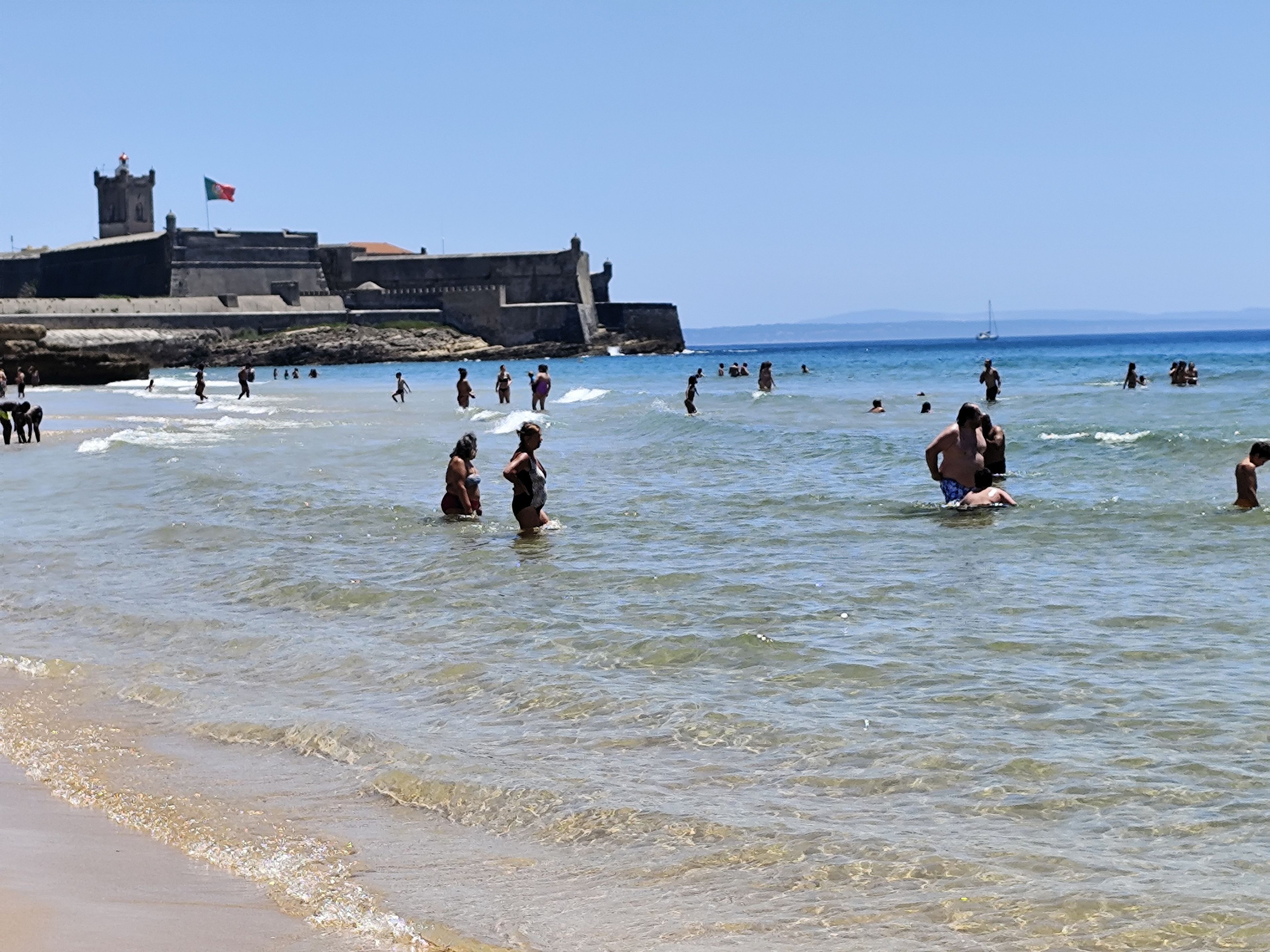 Una playa con gente nadando y caminando por la orilla, un edificio imponente en el fondo y un cielo despejado.