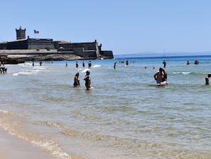 Une plage où l’on voit des gens se baigner et se promener le long du rivage, un grand bâtiment imposant, semblable à une forteresse, en arrière-plan, et un ciel dégagé.