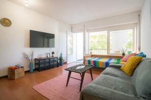 A living room with a sofa, a coffee table, a TV, and a window with a view of greenery.