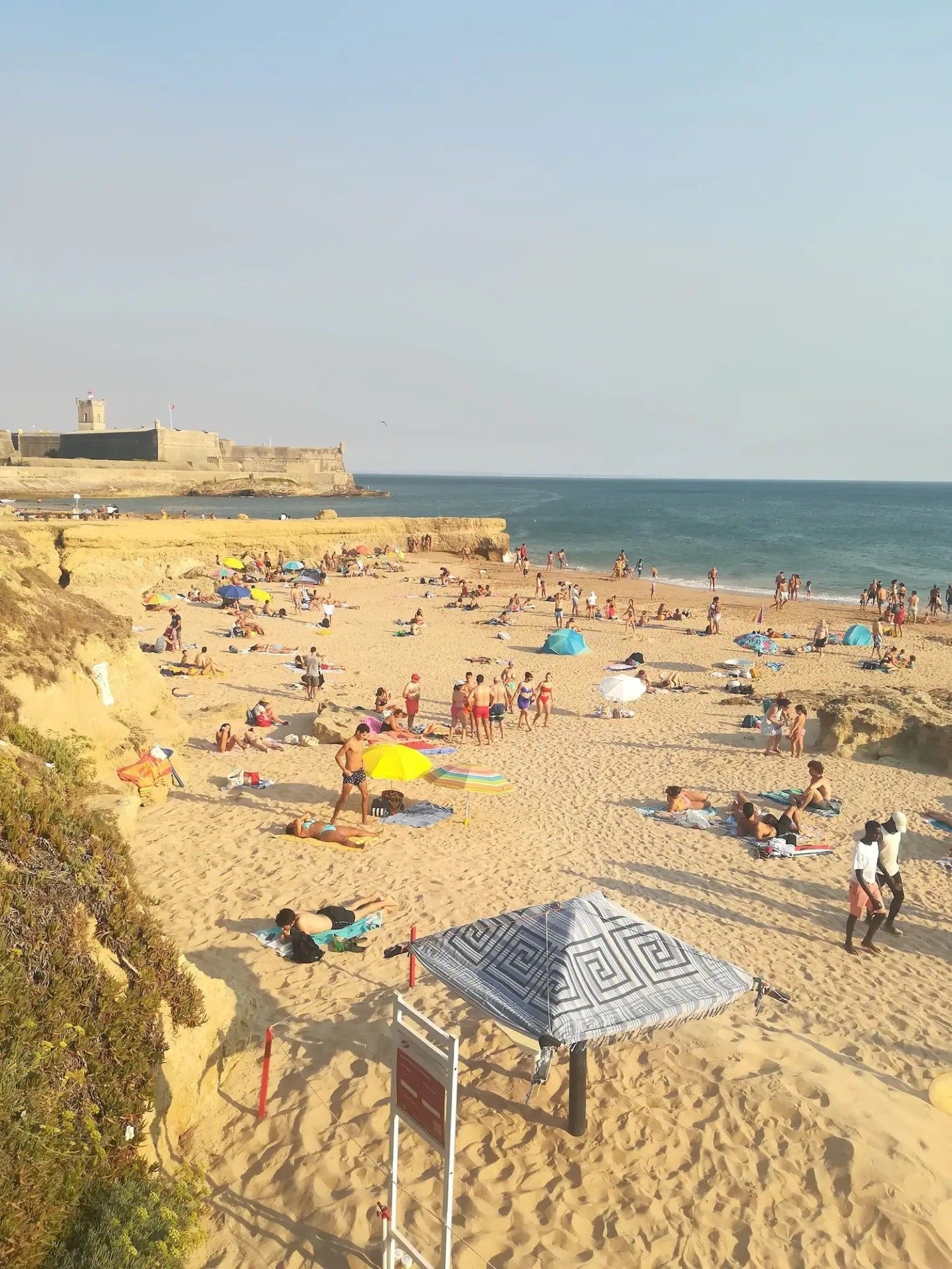 Una playa concurrida, gente tomando el sol y nadando, una orilla arenosa y una fortificación distante.