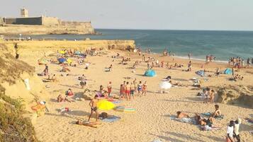 Una playa concurrida, gente tomando el sol y nadando, una orilla arenosa y una fortificación distante.