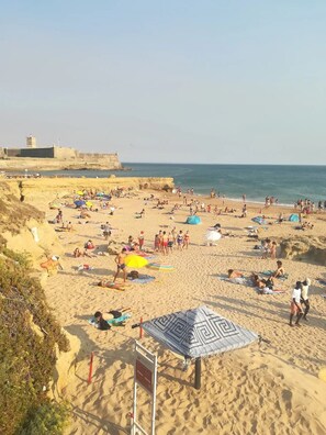 Une plage bondée où les gens se prélassent au soleil et se baignent, une rive sablonneuse et, au loin, une fortification.