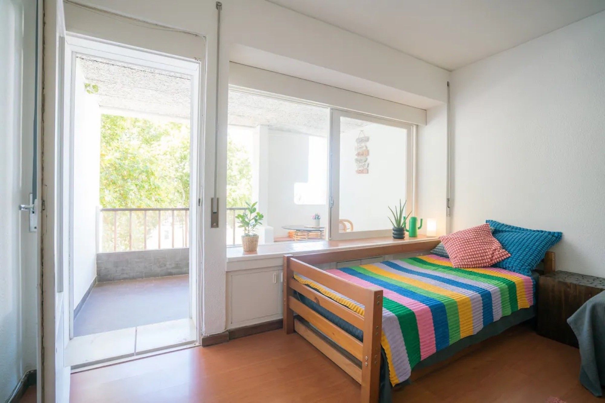 Una habitación pequeña y luminosa con una cama de madera, una ventana con vistas a la vegetación y una puerta que da al exterior.