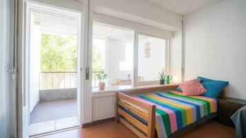 Una habitación pequeña y luminosa con una cama de madera, una ventana con vistas a la vegetación y una puerta que da al exterior.