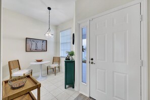 A hallway with a white door, a dining table with chairs, a hanging light fixture, and a framed picture on the wall. - Golfer’s Paradise: Turnkey Rental with 24/7 Fitness & Premier Amenities (Naples)