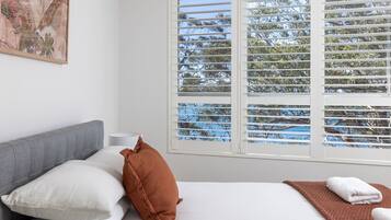 Un dormitorio con cama, almohadas, una mesita de noche y una ventana con persianas.