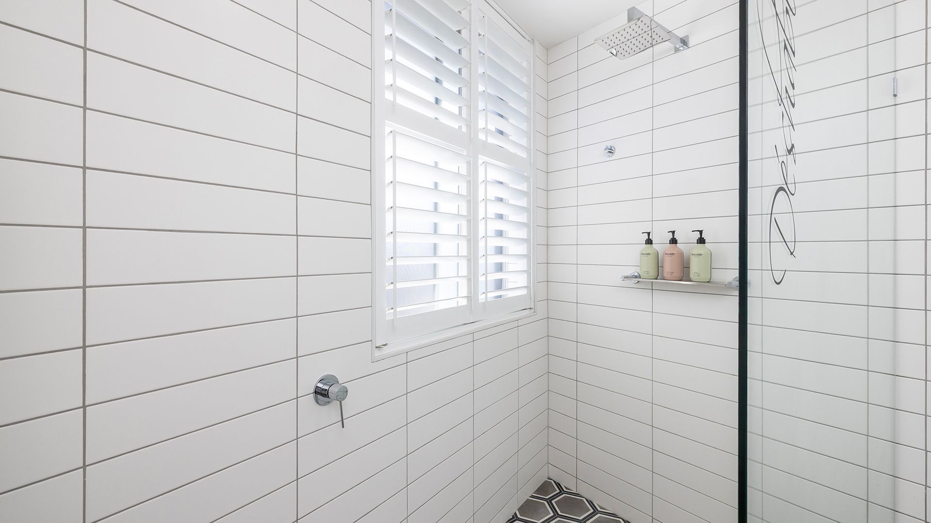 A modern bathroom with white tiled walls, a shower with a glass door, and a window with blinds.